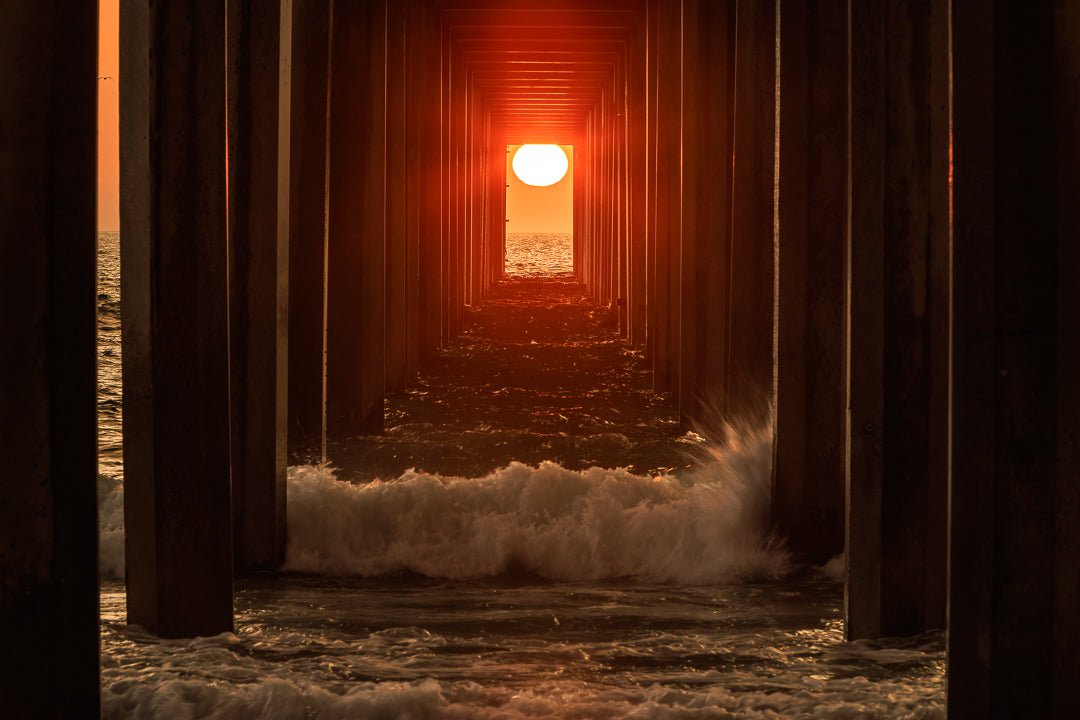Setting sun aligned beneath Scripps Pier during Scrippshenge as waves surge through the illuminated pilings in La Jolla, California